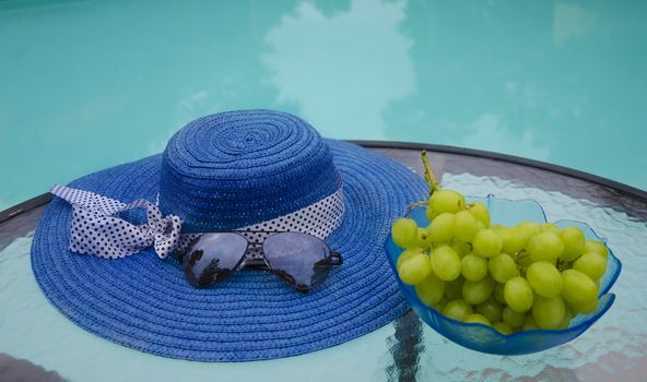 Woman's hat, sunglasses and grape in glass plate are on table by the swimming pool