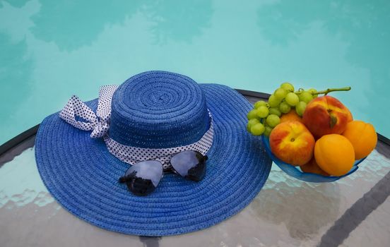 Woman's hat, sunglasses and grape with peaches and apricots in glass plate are on table by the swimming pool