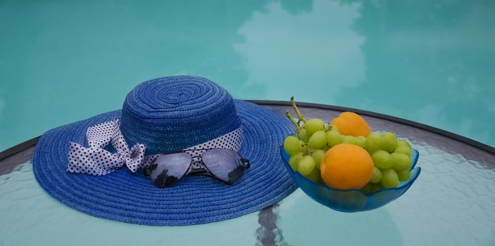 Woman's hat, sunglasses and grape with apricots in glass plate are on table by the swimming pool