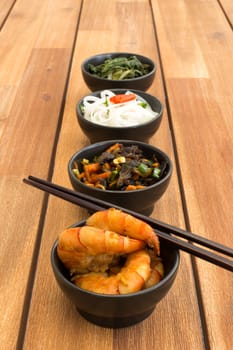 Japanese food composed with four black bowls with shrimps, rice noodles, kale (green cabbage), fried vegetables and asian chopsticks. Composition on a old styled wooden table.