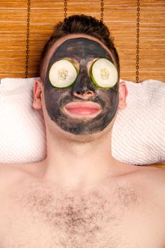 Man face with beauty treatment skincare mask and cucumber laying on bamboo at spa.