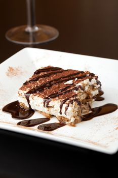 Slice of delicious tirimisu cake on a white plate garnished with cocoa power and drizzled chocolate sauce.