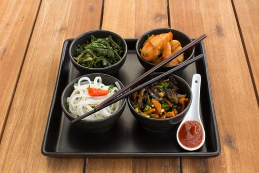 Asian dish on a square plate in black bowls with shrimp, rice noodles, kale (green cabbage) and fried vegetables. Composed with ceramic spoon with spicy red sauce and chinese chopsticks. Composition on a old styled wooden table.