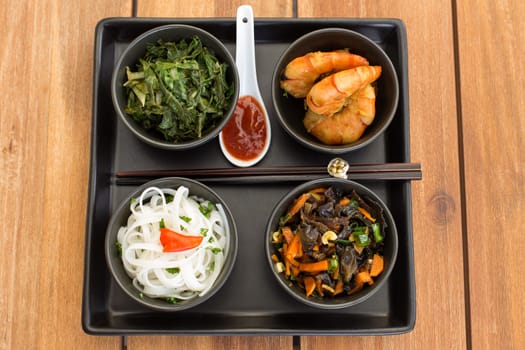 Traditional asian dish on a square plate in black bowls with shrimp, rice noodles, kale (green cabbage) and fried vegetables. Composed with ceramic spoon with spicy red sauce and chinese chopsticks. Composition on a old styled wooden table.