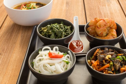 Asian dish on a square plate in black bowls with shrimp, rice noodles, kale (green cabbage) and fried vegetables. Composed with ceramic spoon with spicy red sauce, japanese chopsticks and a bowl of asian soup. Composition on a old wooden table.