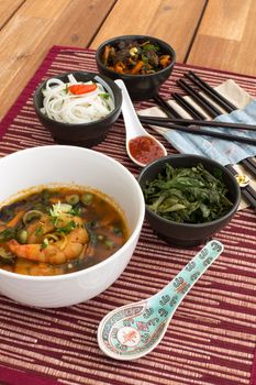 Japanese soup with shrimps in white ceramic bowl composed with ceramic spoon with spicy red sauce, japanese chopsticks and black bowls with rice noodles, kale (green cabbage) and fried vegetables. Composition on a old styled wooden table.