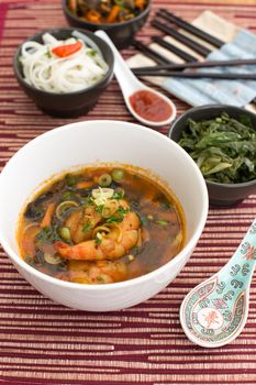 Chinese soup with shrimps in white ceramic bowl composed with ceramic spoon with spicy red sauce, asian chopsticks and black bowls with rice noodles, kale (green cabbage) and fried vegetables.