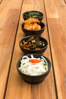 Asian food composed with four black bowls with shrimps, rice noodles, kale (green cabbage) and fried vegetables. Composition on a old styled wooden table.