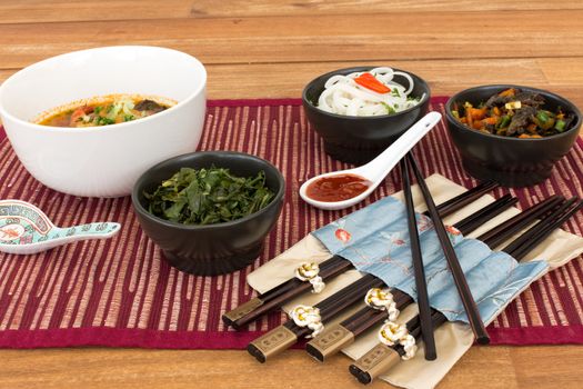 Asian food composed with asian soup with shrimp, ceramic spoon with spicy red sauce, set of asian chopsticks and black bowls with rice noodles, kale (green cabbage) and fried vegetables. Composition on a old styled wooden table.