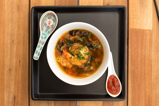 Asian vegetarian soup with shrimps and spicy sauce composed with two ceramic spoons on a square black plate. Composition on a old styled wooden table.