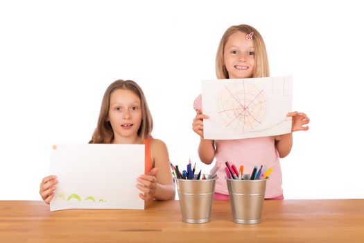 Two sisters with blond hair smiling and showing their super drawings at a wooden table with metallic pencils holder. Isolated on white background.