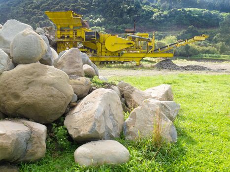 Large boulders and heavy-duty yellow rock crusher machinery used to crush rock in a stone quarry pit or mining site