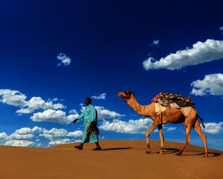 Rajasthan travel background - Indian cameleer (camel driver) with camels in dunes of Thar desert. Jaisalmer, Rajasthan, India