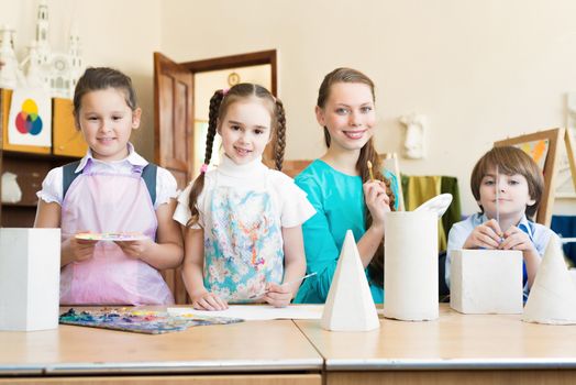 children with the teacher standing at the table, smiling and looking into the cam