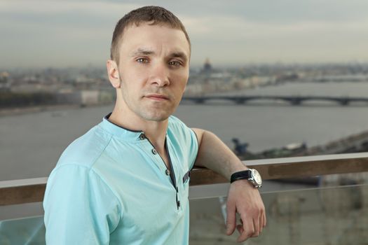 A young man 27 years old, Caucasian, Slav, Russian, dressed in a turquoise shirt with short sleeves, horizontal portrait, close-up, against the background of a panorama of European cities. One person, a male, short hair, outdoor.