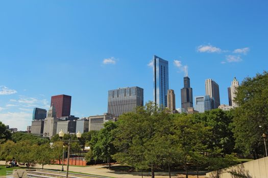 Skyscrapers line the edge of Millennium Park near Columbus Drive to form a partial skyline of Chicago, Illinois, against a bright blue sky with white clouds and space for copy