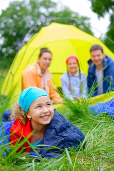 Young girl in a sleeping bag with her family near tent in camping on the nature. Vertical view