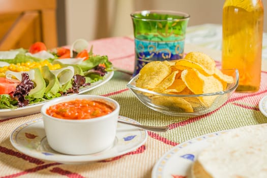 Closeup of traditional mexican food in a table, with  a bowl of nachos, spicy sauce, a plate of tortillas and fresh salad