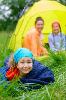 Young girl in a sleeping bag with her family near tent in camping on the nature. Vertical view