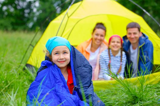 Young girl in a sleeping bag with her family near tent in camping on the nature