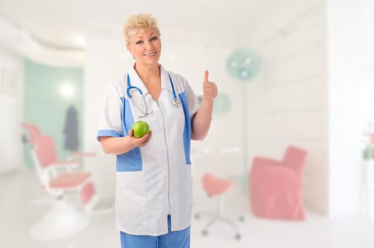 Mature doctor with green apple shows ok gesture at medical office