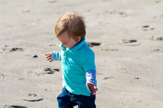 A one year old boy plays at the beach along the coast in the sand while exploring and looking for interesting things.