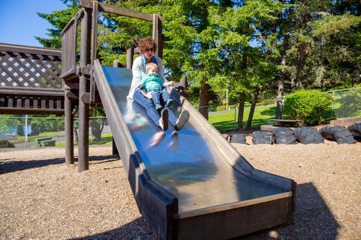 A mom and her son slide down a metal slide at an outdoor playground at a park.