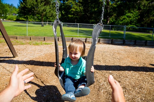 A father pushes his son on a unique looking swing. This image is from the viewpoint of the dad looking towards his son on the swings.