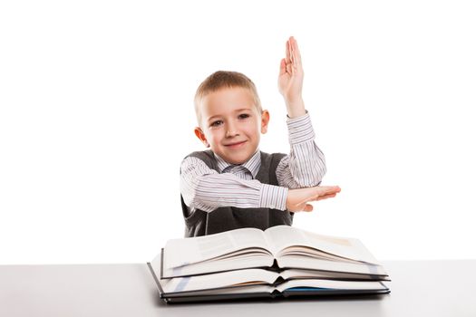 Little smiling child boy with education books at desk gesturing hand up for answering school homework lesson