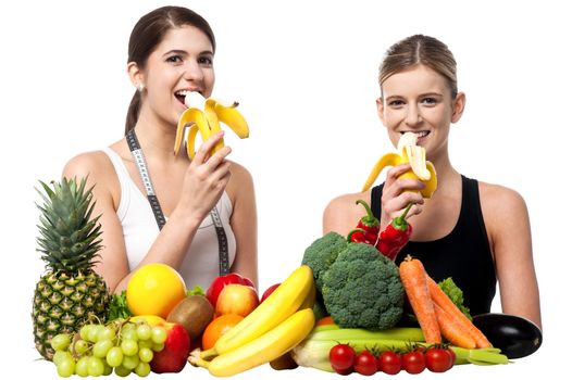 Two pretty girls, each eating banana. Fruits all around them