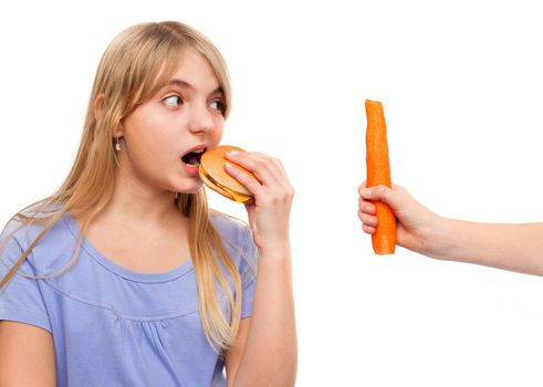 Young female eating a cheeseburger looking at a hand holding a carrot. Isolated on white.