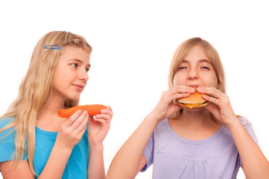 Girl eating a carrot looking at another girl sadly who is eating a hamburger. Isolated on white.