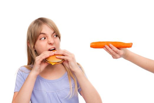 Dilemma of healthy eating - Girl eating a cheeseburger looks at a hand holding a carrot. Isolated on white