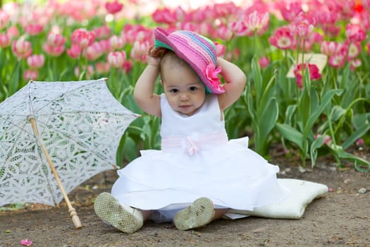 baby in an elegant dress with an umbrella sits near blossoming tulips