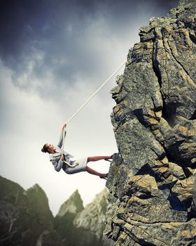 businesswoman climbing steep mountain hanging on rope