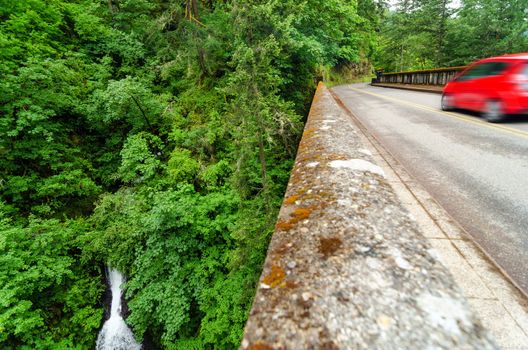 Waterfall in a lush green forest in Oregon next to historic Highway 30 with a red blurry car