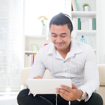 Handsome Asian man using tablet computer. Smiling Southeast Asian college student relaxing and listening to music at home. Asian model.