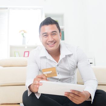 Attractive Southeast Asian man using a credit card to shop on the internet with a tablet computer. Asian male model at home.