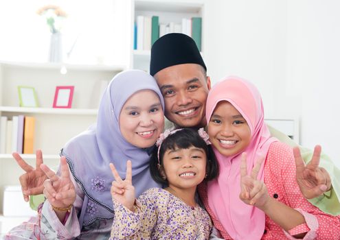 Happy Asian family at home. Muslim family showing v victory hand sign and having fun. Southeast Asian parents and children smiling.