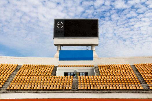 Rows of orange seats on the stadium with scoreboard displaying clock above them