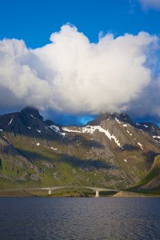 Bridge connecting islands on Norwegian coast with high mountains, patches of snow and peaks in the clouds