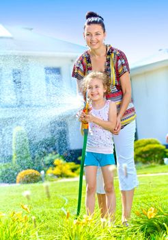 Little gardener girl with mother watering flowers on the lawn near cottage. Vertical view