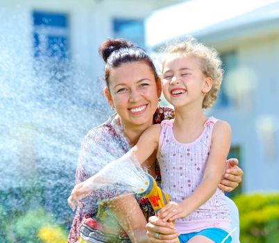 Portrait of little gardener girl with mother watering flowers on the lawn near cottage.