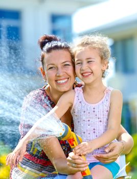 Portrait of little gardener girl with mother watering flowers on the lawn near cottage. Vertical view