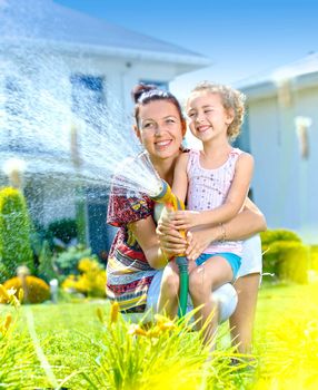 Little gardener girl with mother watering flowers on the lawn near cottage. Vertical view