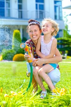 Little gardener girl with mother watering flowers on the lawn near cottage. Vertical view