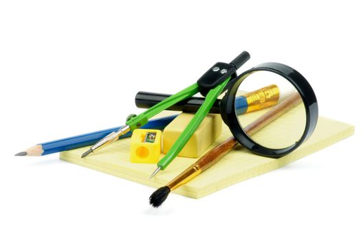 Arrangement of Pencil, Pen, Paintbrush, Pencil Sharpener, Eraser, Drawing Compass and Magnifier on Yellow Note Pad isolated on white background