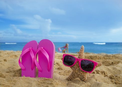 Girl's Flip flops and starfish with sunglasses on sandy beach with playing girl by the ocean on the background in Hawaii