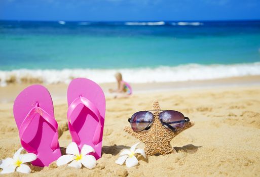Flip flops and starfish with sunglasses with tropical flowers on sandy beach with playing girl by the sea on background (Hawaii, Kauai)