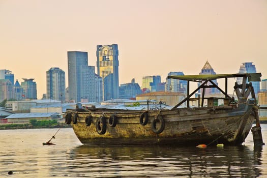 Old disfunctioned boat at sunset on the Chao Phraya river in the outskirts of Bangkok, Tahiland with panorama of Silom district